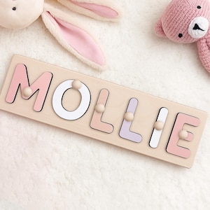 May include: A wooden name puzzle spelling "MOLLIE" in pastel pink, white, and lavender letters. Each letter has a wooden peg for easy handling. A plush bunny and a pink teddy bear are in the background.