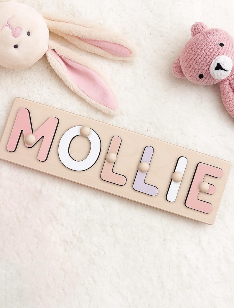 May include: A wooden name puzzle spelling "MOLLIE" in pastel pink, white, and lavender letters. Each letter has a wooden peg for easy handling. A plush bunny and a pink teddy bear are in the background.