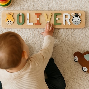 May include: Wooden name puzzle spelling "OLIVER" with colorful letters and animal designs. The letters are in green, white, orange, and yellow. A giraffe and zebra are on either end of the puzzle. A child's hand is reaching for the puzzle.