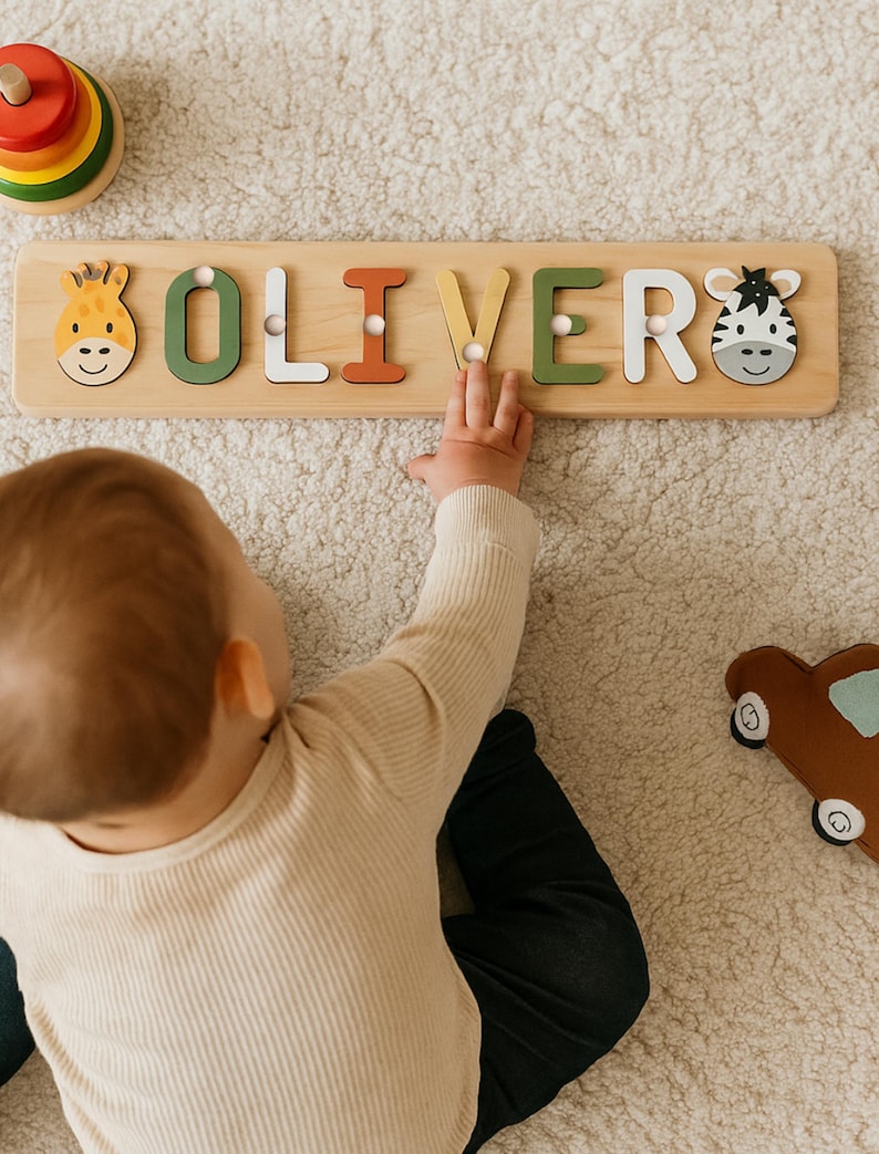 May include: Wooden name puzzle spelling "OLIVER" with colorful letters and animal designs. The letters are in green, white, orange, and yellow. A giraffe and zebra are on either end of the puzzle. A child's hand is reaching for the puzzle.
