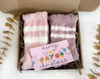 May include: A gift box with two pairs of pink and white striped fuzzy socks and a pink card that says "merry christmas" with colorful holiday icons.