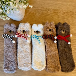 May include: Five pairs of fuzzy bear-shaped socks in various shades of brown and white. Each sock has a different colored scarf around the neck, including black and white stripes, red and white, and blue.