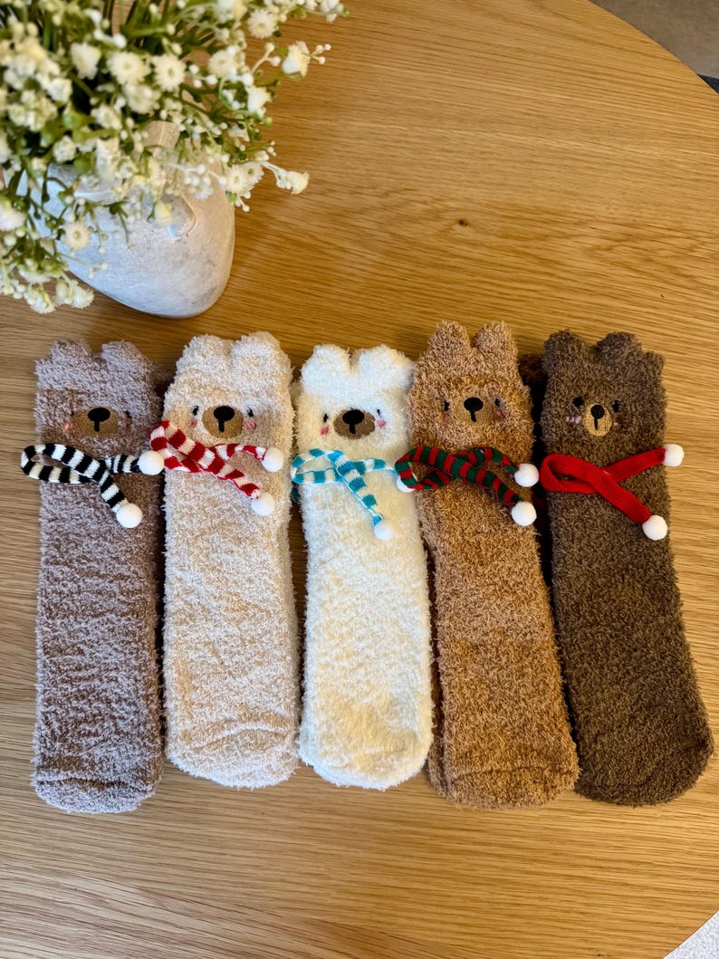 May include: Five pairs of fuzzy bear-shaped socks in various shades of brown and white. Each sock has a different colored scarf around the neck, including black and white stripes, red and white, and blue.