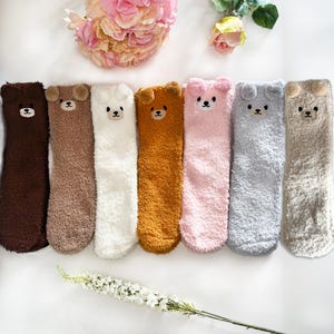 May include: Seven pairs of fuzzy crew socks in dark brown, light brown, white, mustard yellow, light pink, light gray, and beige. Each sock features a plush bear face at the top with small rounded ears, embroidered black eyes, and a nose.