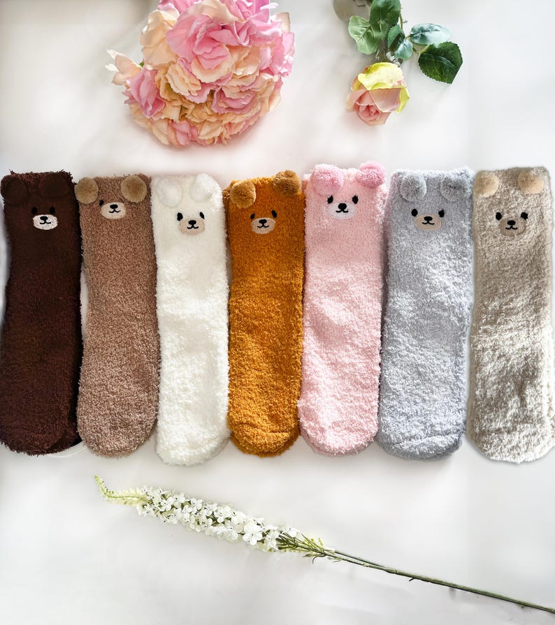 May include: Seven pairs of fuzzy crew socks in dark brown, light brown, white, mustard yellow, light pink, light gray, and beige. Each sock features a plush bear face at the top with small rounded ears, embroidered black eyes, and a nose.