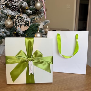 May include: A square white gift box with a textured surface, wrapped in a vibrant lime green satin ribbon tied into a large bow. A small white rectangular tag attached to the ribbon features the printed text Best wishes in black ink.
