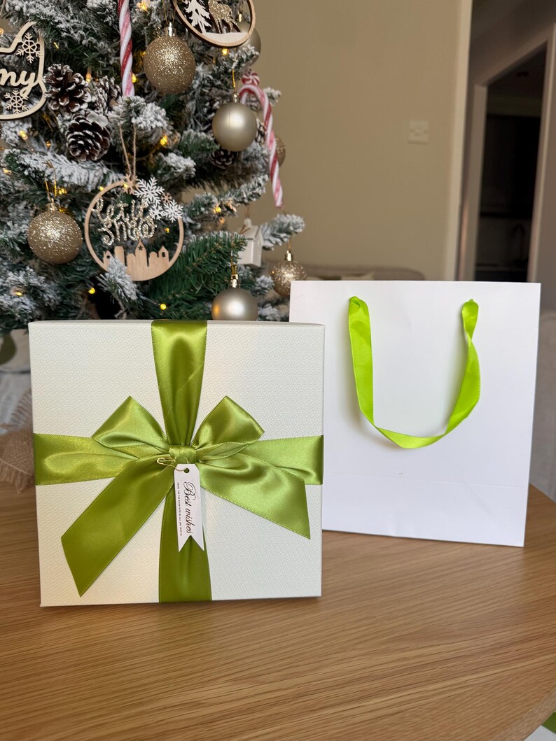 May include: A square white gift box with a textured surface, wrapped in a vibrant lime green satin ribbon tied into a large bow. A small white rectangular tag attached to the ribbon features the printed text Best wishes in black ink.
