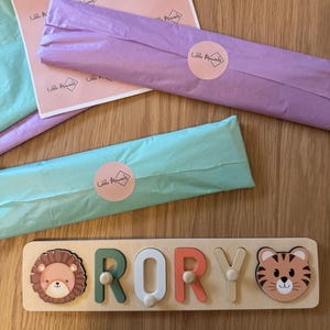 May include: Wooden name puzzle spelling "RORY" with a lion and tiger design. The letters are in green, white, and coral. The puzzle is on a wooden surface, with wrapped packages in pink and teal tissue paper.