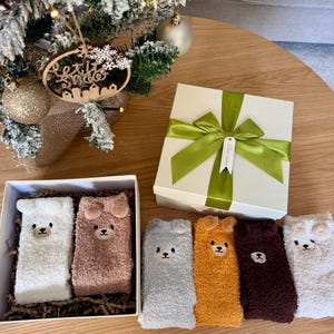 May include: A set of six fuzzy crew socks featuring embroidered bear faces with small ears. The socks come in white, light brown, gray, mustard yellow, dark brown, and cream. Two socks are inside a white gift box, and four are arranged outside.
