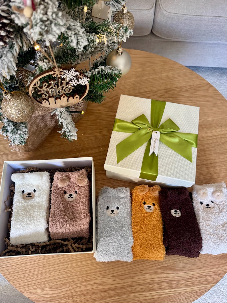 May include: A set of six fuzzy crew socks featuring embroidered bear faces with small ears. The socks come in white, light brown, gray, mustard yellow, dark brown, and cream. Two socks are inside a white gift box, and four are arranged outside.