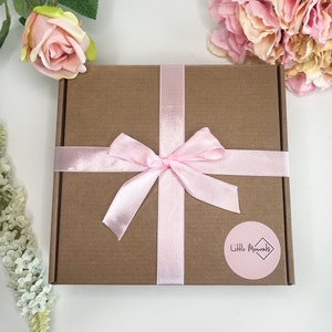 May include: A square brown cardboard gift box tied with a light pink satin ribbon in a bow. A round pink sticker on the box features the text Little Moments inside a diamond shape. The box is surrounded by artificial pink and white flowers.
