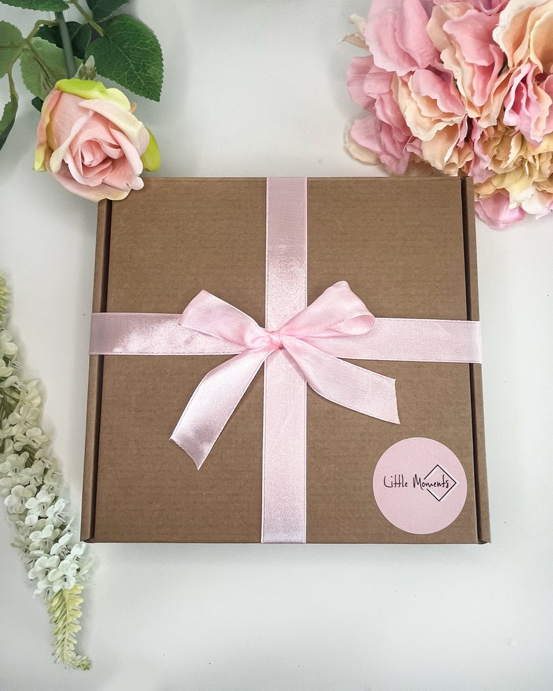 May include: A square brown cardboard gift box tied with a light pink satin ribbon in a bow. A round pink sticker on the box features the text Little Moments inside a diamond shape. The box is surrounded by artificial pink and white flowers.