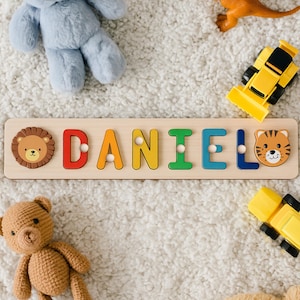 May include: Wooden name puzzle spelling "DANIEL" with colorful letters and animal-themed end pieces. The puzzle is surrounded by toy cars, a teddy bear, a plush bear, and a dinosaur toy on a white, fluffy surface.