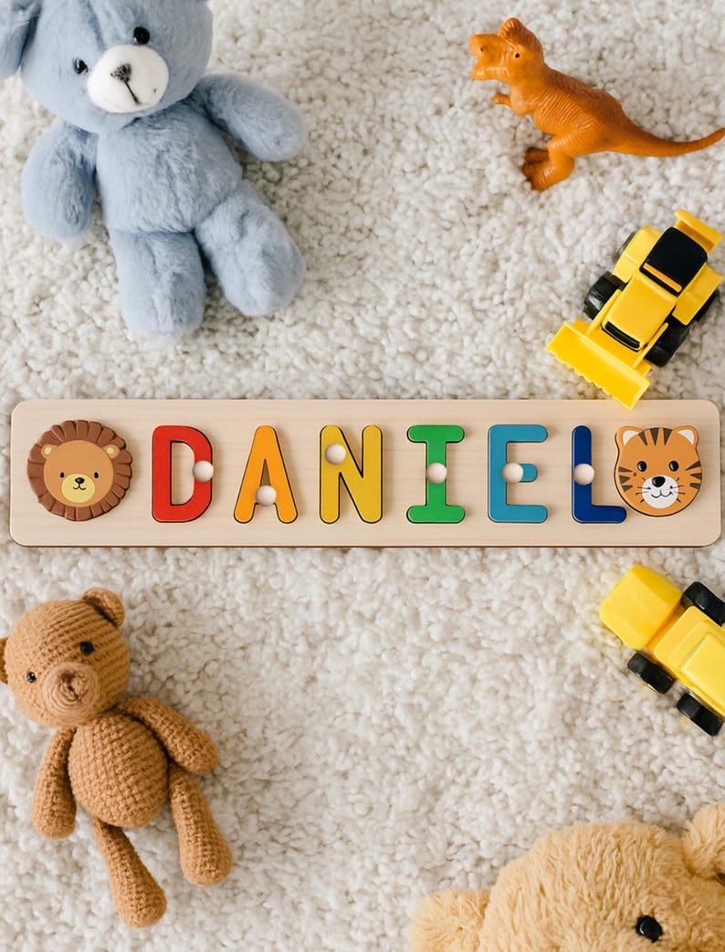 May include: Wooden name puzzle spelling "DANIEL" with colorful letters and animal-themed end pieces. The puzzle is surrounded by toy cars, a teddy bear, a plush bear, and a dinosaur toy on a white, fluffy surface.