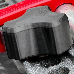 Puede incluir: Primer plano de un pomo negro texturizado con una parte superior en forma de estrella, asegurado por una abrazadera roja. El pomo está sujeto a una barra de metal y la abrazadera a un marco metálico.