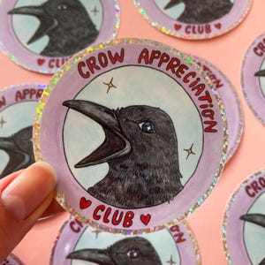 Könnte beinhalten: Eine Sammlung runder Aufkleber mit einer Krähenillustration. Jeder Aufkleber zeigt eine schwarze Krähe mit offenem Schnabel, umgeben von dem Text "CROW APPRECIATION CLUB". Die Aufkleber haben einen hellvioletten Rand mit holografischem Glitzereffekt.
