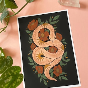 May include: A print featuring a coiled snake illustration. The snake is a light peach colour with starburst details, surrounded by orange flowers and green leaves on a black background. The artwork is on a white paper.