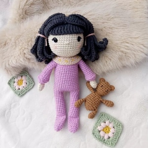 May include: A crocheted doll with black hair and a purple outfit is holding a brown crocheted teddy bear. The doll and bear are on a white fluffy surface with two green and pink crocheted squares with flower designs.