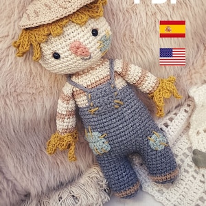 May include: A crocheted scarecrow doll with a straw hat, overalls, and a friendly face. The doll is lying on a fluffy, light brown surface. The image also includes the text "PDF" and flags for Spain and the United States.