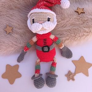 Puede incluir: Un muñeco de crochet de Papá Noel con un gorro rojo, barba blanca y piernas a rayas rojas y verdes. El muñeco está sentado sobre una superficie blanca con estrellas marrones.