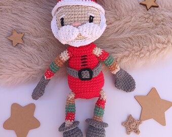 Puede incluir: Un muñeco de crochet de Papá Noel con un gorro rojo, barba blanca y piernas a rayas rojas y verdes. El muñeco está sentado sobre una superficie blanca con estrellas marrones.