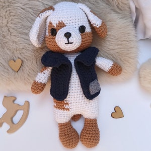 Puede incluir: Un juguete de peluche de perro de crochet con pelo marrón y blanco, que lleva un chaleco azul. El perro tiene ojos marrones y una nariz negra. El juguete está sentado sobre una superficie blanca.