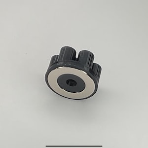 Puede incluir: Una perilla de plástico negro con un anillo de metal plateado alrededor del centro. La perilla tiene múltiples perillas pequeñas alrededor del borde.