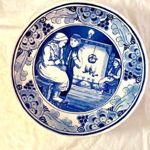 May include: A blue and white ceramic plate with a Dutch Delft design. The plate features a scene of three people in traditional clothing standing in front of a fireplace. The plate is decorated with floral motifs and a border of blue and white.