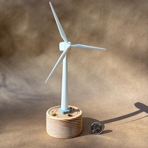 May include: A miniature white wind turbine model with three blades, mounted on a wooden base. The base has a light brown finish and is filled with sand and small tufts of grass. A quarter coin is next to the model.