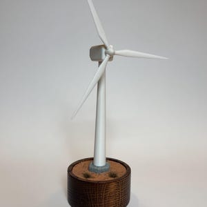 May include: A white wind turbine model with three blades mounted on a cylindrical wooden base. The base features a textured tan surface with small patches of green grass and a gray stone-like foundation for the turbine tower.