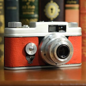May include: A vintage Argus camera with a red leatherette body and a white top. The camera has a silver lens and dials, with the Argus logo visible. The camera is set against a backdrop of antique books.