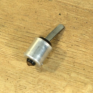 Puede incluir: Una pequeña herramienta plateada con un cuerpo cilíndrico y una hoja rectangular plana. La herramienta tiene una base negra y está colocada sobre una superficie de madera. La herramienta parece ser un instrumento especializado, posiblemente para manualidades o reparación.