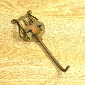 May include: A vintage brass music lyre clip. The clip has a lyre-shaped top with a central design and a long, slender stem ending in a hook. The brass has a warm, aged patina, suggesting its age. The clip is designed to hold sheet music.