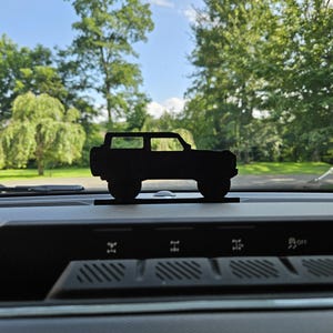 Peut inclure: Silhouette noire d'un SUV à quatre portes posée sur un tableau de bord. Le véhicule est une découpe détaillée, mettant en valeur le style de la carrosserie. L'arrière-plan comprend un ciel bleu et des arbres verts.