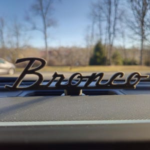 May include: A dark brown "Bronco" emblem, mounted on a textured dashboard. The emblem is in a cursive font. The background shows a car, trees, and a clear blue sky.