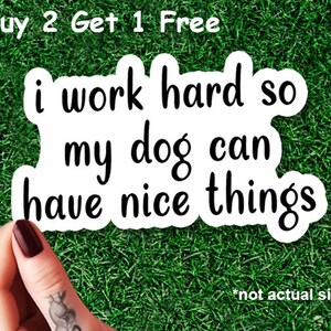Könnte beinhalten: Weißer Aufkleber mit dem Text "i work hard so my dog can have nice things" in schwarzer, handschriftlicher Schrift. Der Aufkleber hat auch den Text "Buy 2 Get 1 Free" und "*not actual size".
