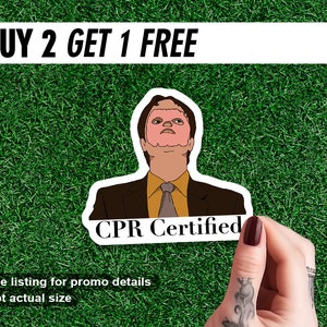 May include: A sticker featuring a cartoon image of a man in a suit with the text "CPR Certified." Above the sticker is a banner that reads "BUY 2 GET 1 FREE." The sticker is held against a green grass background.