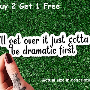 May include: White sticker with the text "I'll get over it just gotta be dramatic first" in a black, bold font. The sticker is held against a green grass background. The text "Buy 2 Get 1 Free" is at the top.