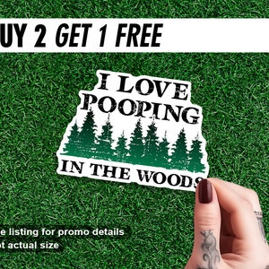 May include: A white sticker with the text "I LOVE POOPING IN THE WOODS" in black and green, with a forest graphic. The words "BUY 2 GET 1 FREE" are at the top. The sticker is on a green grassy surface.