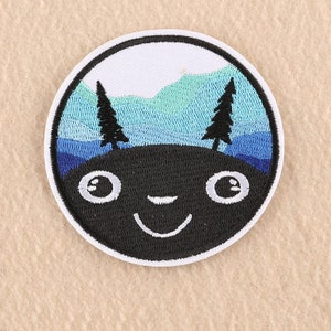 May include: A circular embroidered patch featuring a black face with large white eyes and a smiling mouth. Above the face, the design depicts a landscape with two black pine trees set against layered blue and light blue mountains and a white sky.