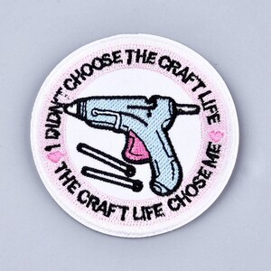 May include: White round embroidered patch with pink lettering that reads "I didn't choose the craft life, the craft life chose me."  A pink and blue glue gun is in the center of the patch with three white glue sticks.