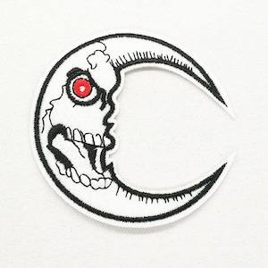 May include: A crescent moon-shaped embroidered patch featuring a white skull design with a single prominent red eye. The patch is outlined in thick black thread, with black stitching detailing the skull's teeth, jaw, and facial features.