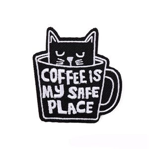 May include: Black and white embroidered patch of a cat inside a coffee mug with the text "Coffee is my safe place".