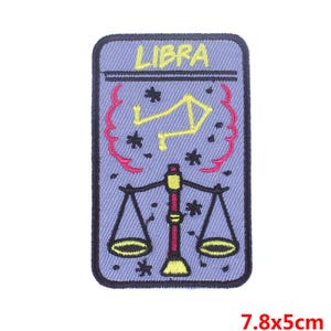 May include: A purple embroidered iron-on patch with the word "LIBRA" in yellow lettering. The patch features a stylized image of a scale with a constellation of stars above it. The patch measures 7.8 by 5 centimeters.