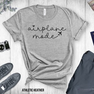 Puede incluir: Una camiseta gris con el texto "airplane mode" en letras negras. La camiseta est&aacute; sobre una superficie de madera blanca con una c&aacute;mara, un pasaporte y un pantal&oacute;n.