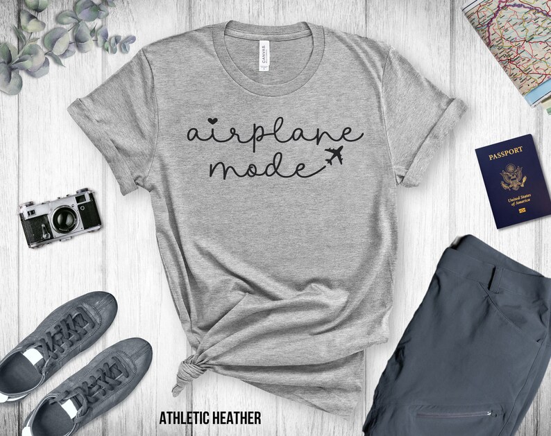 Puede incluir: Una camiseta gris con el texto "airplane mode" en letras negras. La camiseta est&aacute; sobre una superficie de madera blanca con una c&aacute;mara, un pasaporte y un pantal&oacute;n.