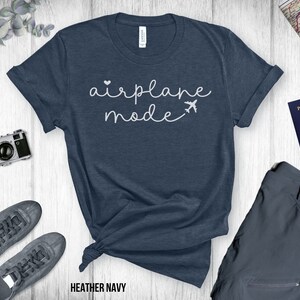 Puede incluir: Una camiseta azul marino jaspeada con el texto "airplane mode" en letras blancas cursivas. Un peque&ntilde;o coraz&oacute;n blanco y un icono de avi&oacute;n est&aacute;n por encima del texto.