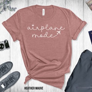 Puede incluir: Una camiseta de color malva con el texto "airplane mode" en letras blancas con un peque&ntilde;o coraz&oacute;n y un gr&aacute;fico de avi&oacute;n.
