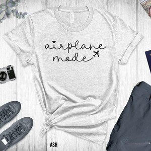 Puede incluir: Una camiseta gris con el texto "airplane mode" en letras negras con un peque&ntilde;o coraz&oacute;n y un gr&aacute;fico de avi&oacute;n. La camiseta est&aacute; colocada sobre una superficie de madera blanca con un pasaporte, una c&aacute;mara y zapatos.