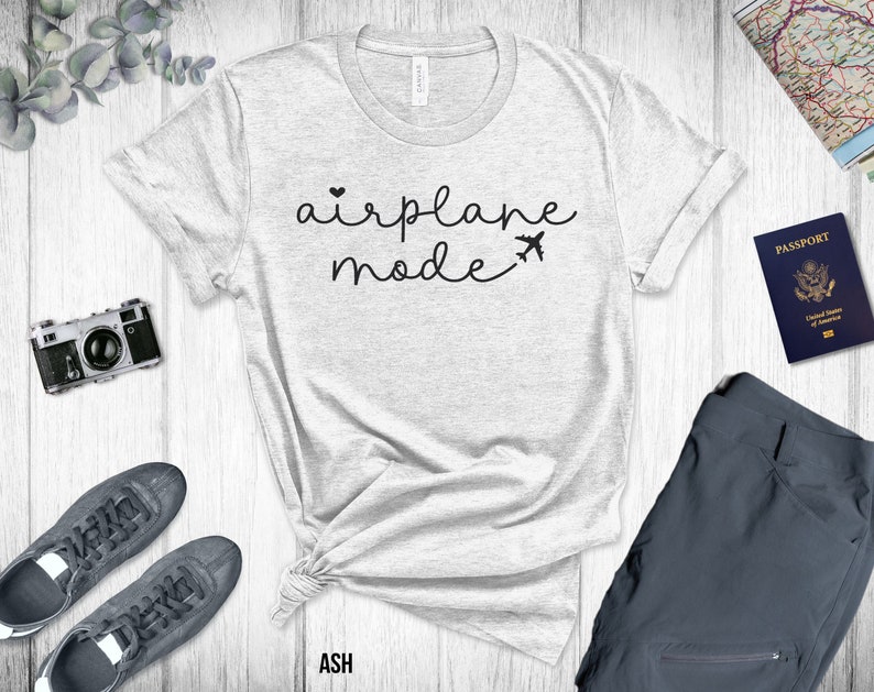 Puede incluir: Una camiseta gris con el texto "airplane mode" en letras negras con un peque&ntilde;o coraz&oacute;n y un gr&aacute;fico de avi&oacute;n. La camiseta est&aacute; colocada sobre una superficie de madera blanca con un pasaporte, una c&aacute;mara y zapatos.